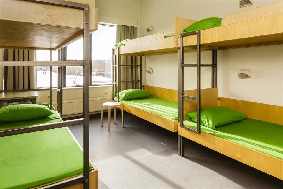 Literas en una habitación bien iluminada con sábanas y almohadas verdes. Un pequeño taburete junto a una ventana.