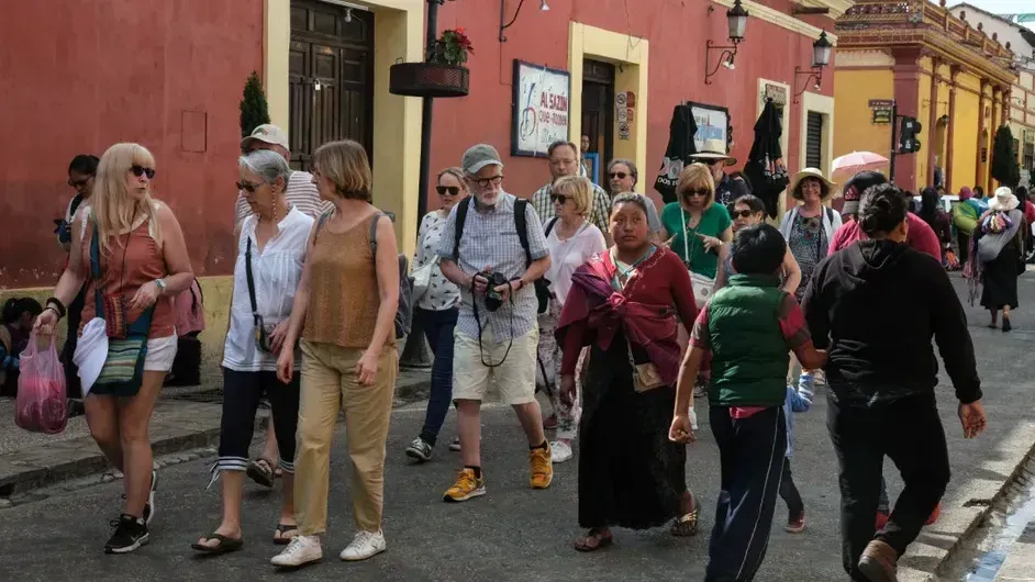 Grupo de personas caminando por una calle de la ciudad; edificios rojos y amarillos en el fondo.