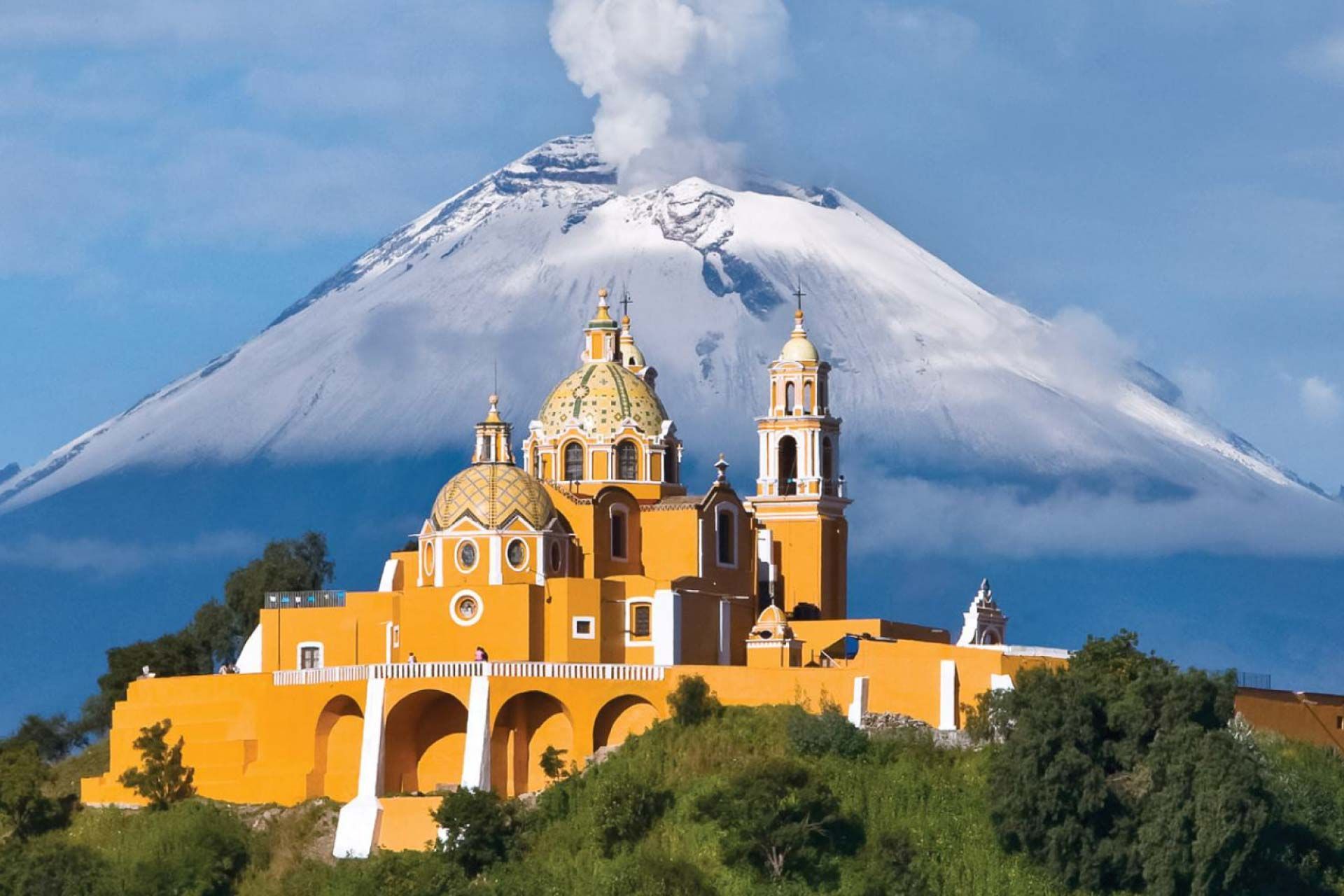 Iglesia amarilla con cúpulas doradas, volcán cubierto de nieve al fondo, cielo azul y colina verde.