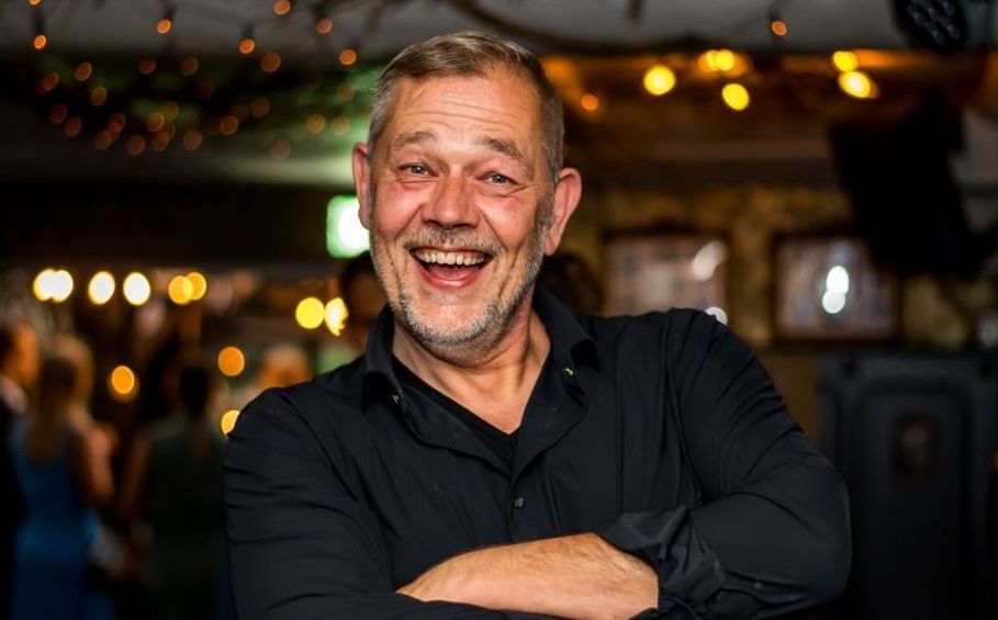 Een man in een zwart shirt lacht, met zijn armen over elkaar, in een sfeervol verlichte bar.