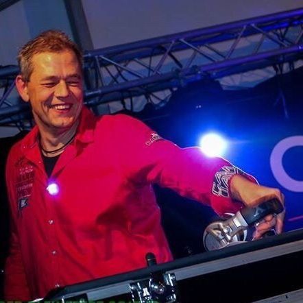 Een man in een rood shirt lacht terwijl hij als dj optreedt op een evenement met felle lichten.