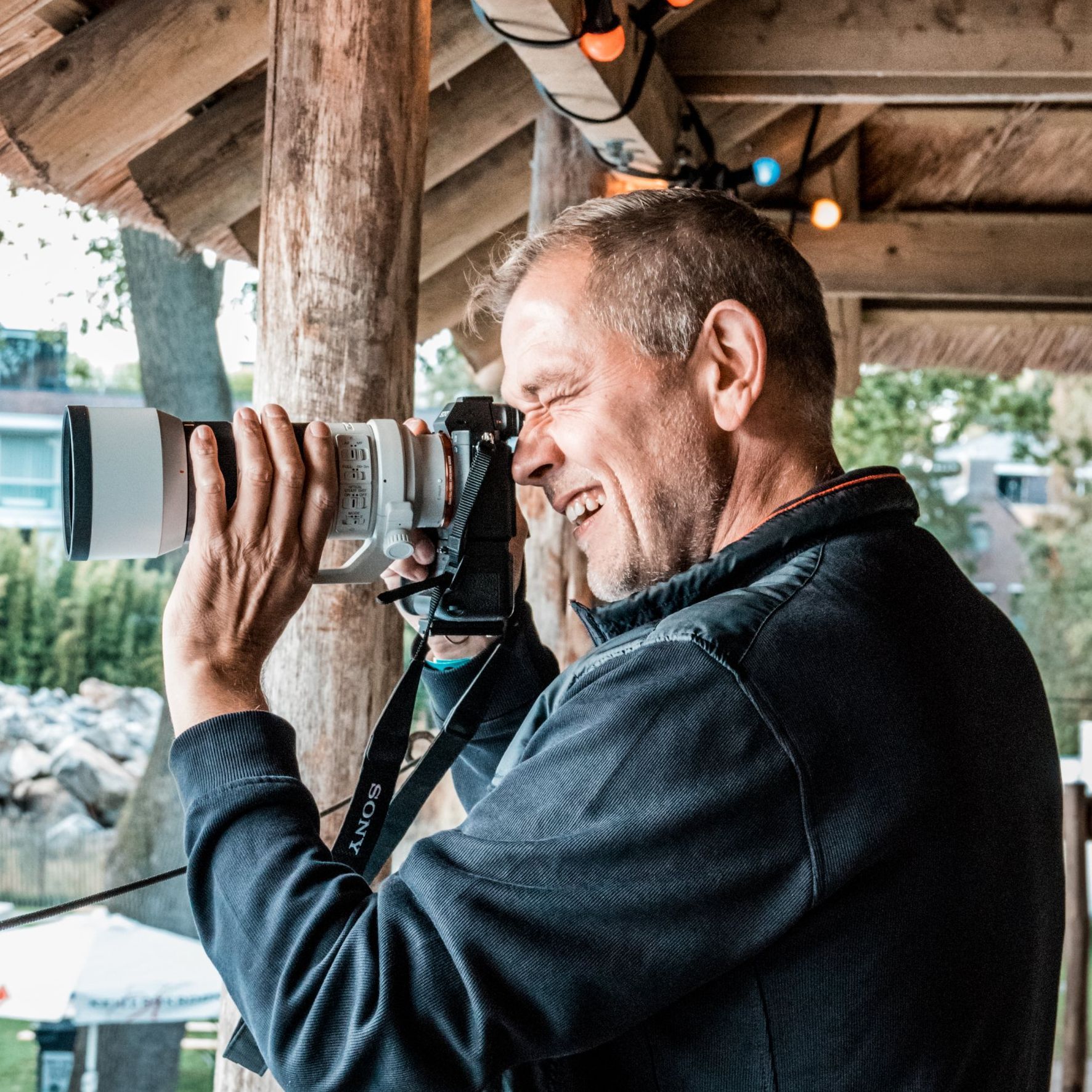 Een glimlachende man kijkt door een camera met een grote lens; buiten, zonnig weer.