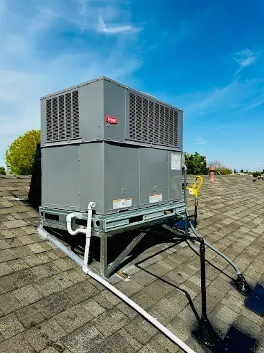 Rooftop HVAC unit on a metal frame against a blue sky. Gray unit, white pipes, black roofing.