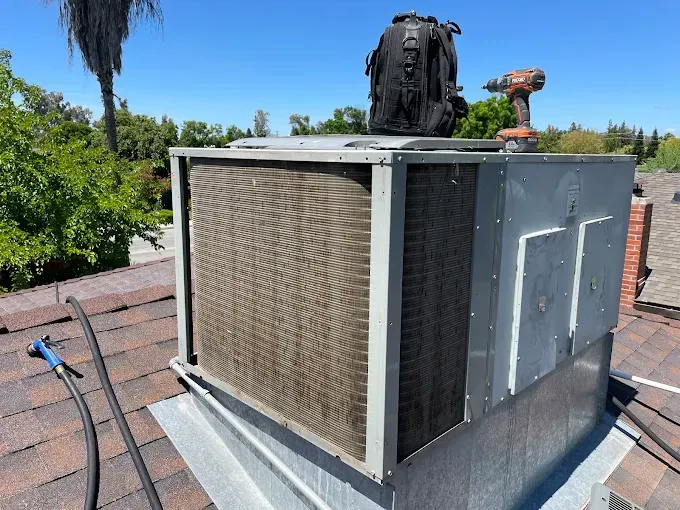 HVAC unit on a rooftop with a backpack and drill. Brown and silver. Sunny day.