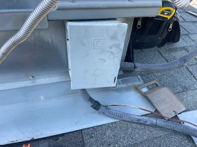 Gray electrical box attached to a metal unit on a rooftop, with conduits and tools visible.
