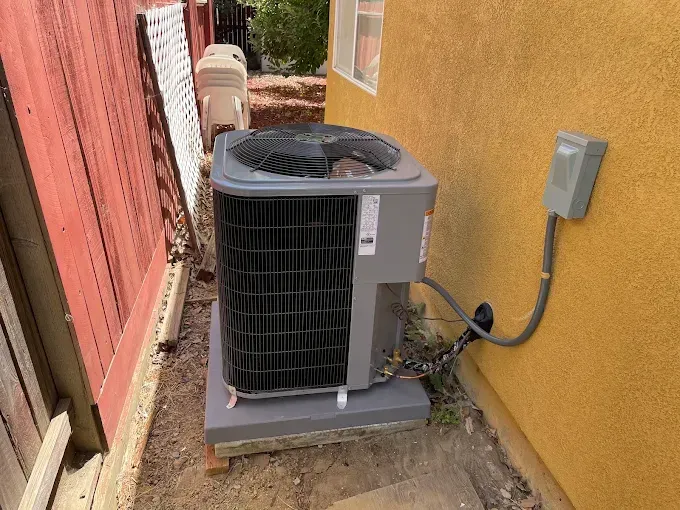 Air conditioning unit next to a wall. Unit is gray and black, sitting on a concrete pad. Electrical box on the wall.