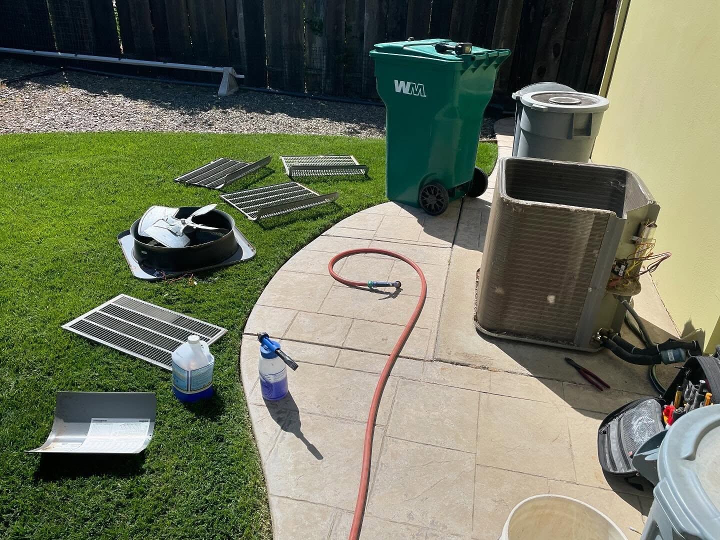 AC unit being cleaned outdoors, various parts laid on grass and patio, hose, cleaning supplies.