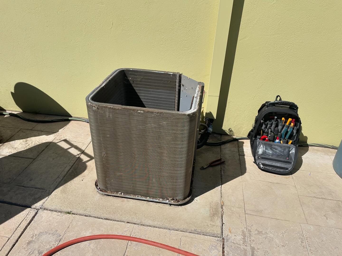 Air conditioning unit coil being worked on next to a tool bag. Outdoor setting.