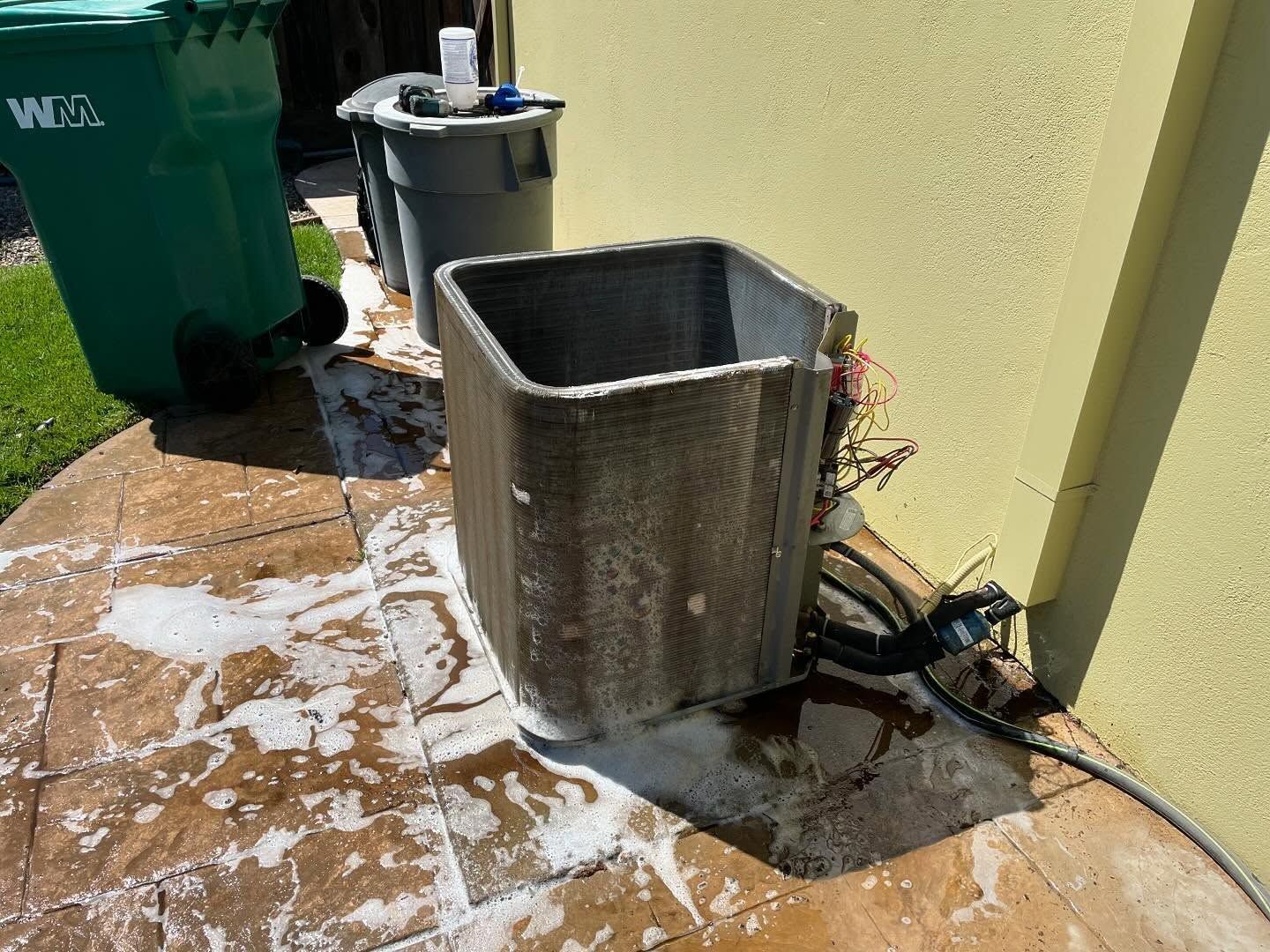 Air conditioner unit being cleaned with soapy water on a paved patio next to a yellow wall and green trash can.