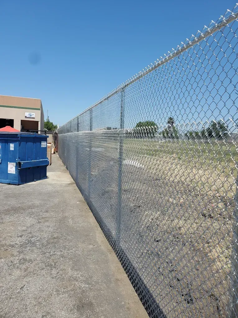 A long chain-link fence stretches into the distance, next to a blue dumpster on a paved lot under a clear blue sky.