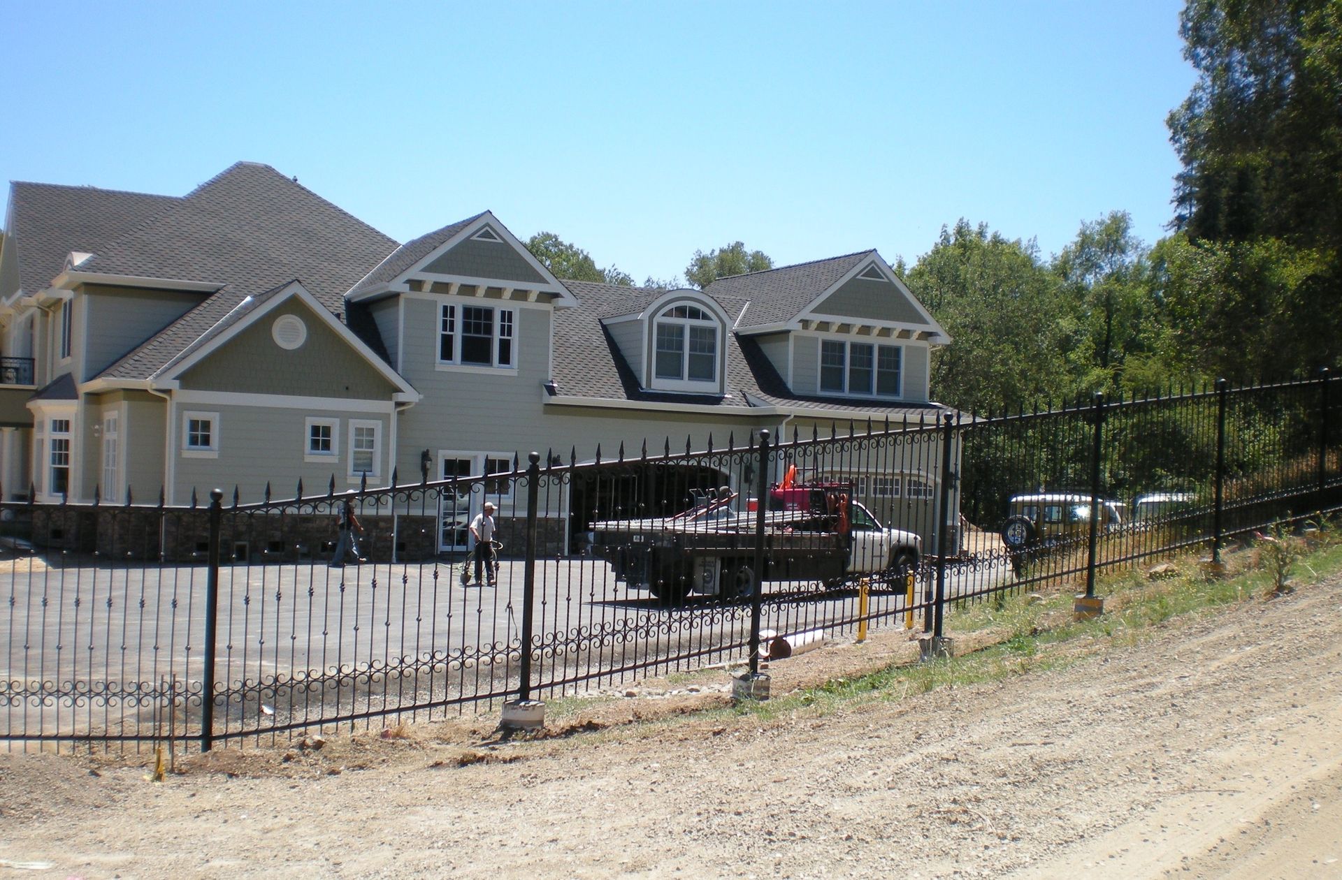 A multi-story house with light siding and grey roofing, viewed from behind a black wrought iron fence on a sunny day.