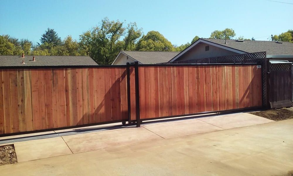 A wooden sliding gate with a black metal frame installed along a concrete driveway in a residential neighborhood.