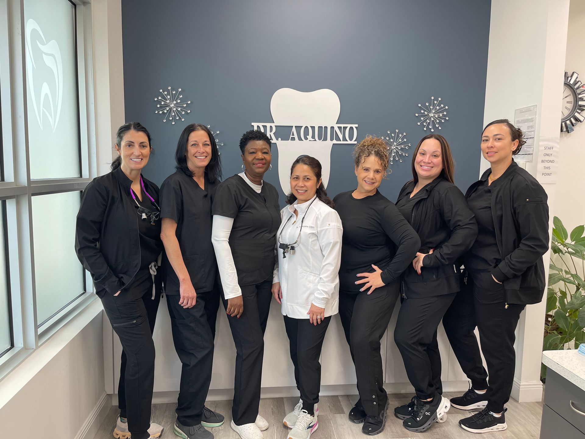 Seven dental staff smiling, posing in front of a logo and window. They are wearing scrubs.