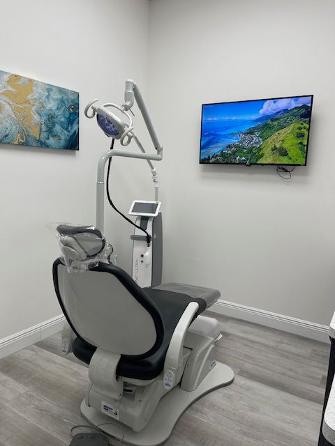 Modern dental treatment room with reclining exam chair, overhead lamp, and wall-mounted TV.