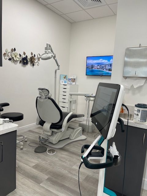 Dental exam room with a reclining chair, overhead light, wall-mounted screen, and equipment cart.