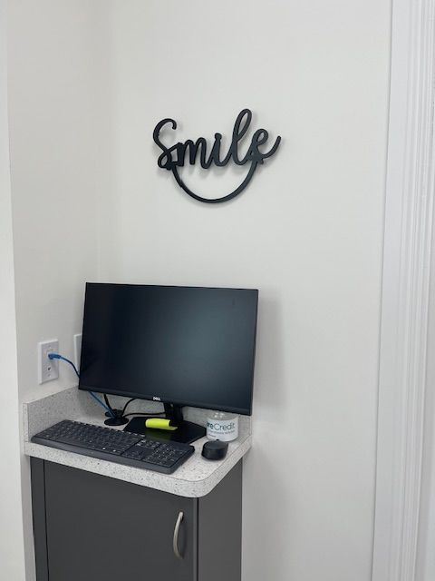 Small office corner with a computer desk, monitor, and a “smile” wall sign.