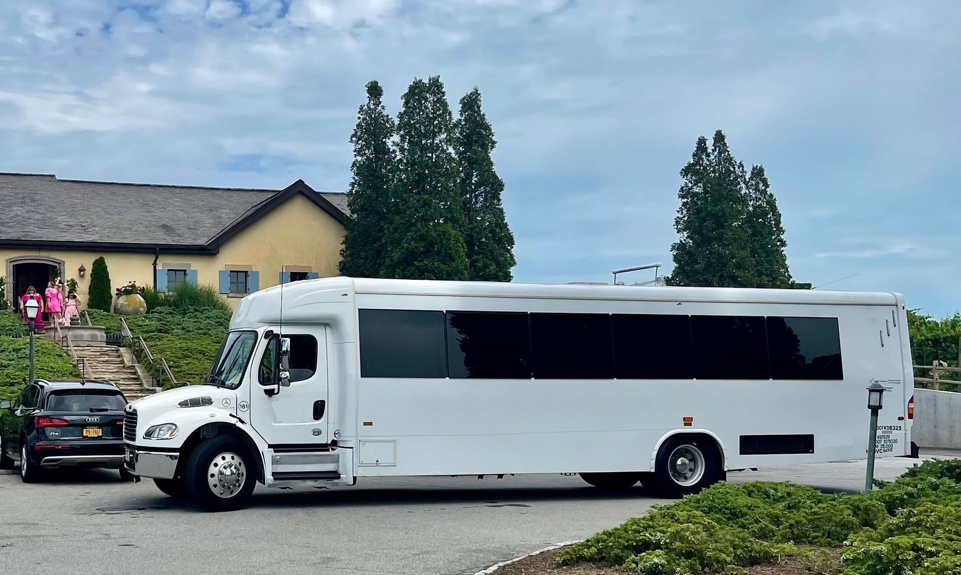 A white party bus parked in a gravel lot in front of a building with a beige stucco exterior and tall pine trees.