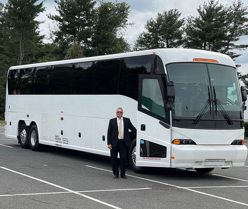 A person in a suit stands next to a white passenger bus in an outdoor parking lot.