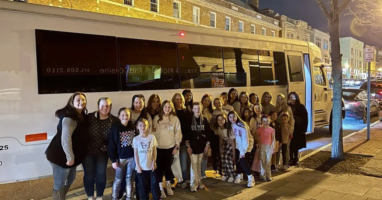 A group stands smiling in front of a white party bus parked on a city street at night.