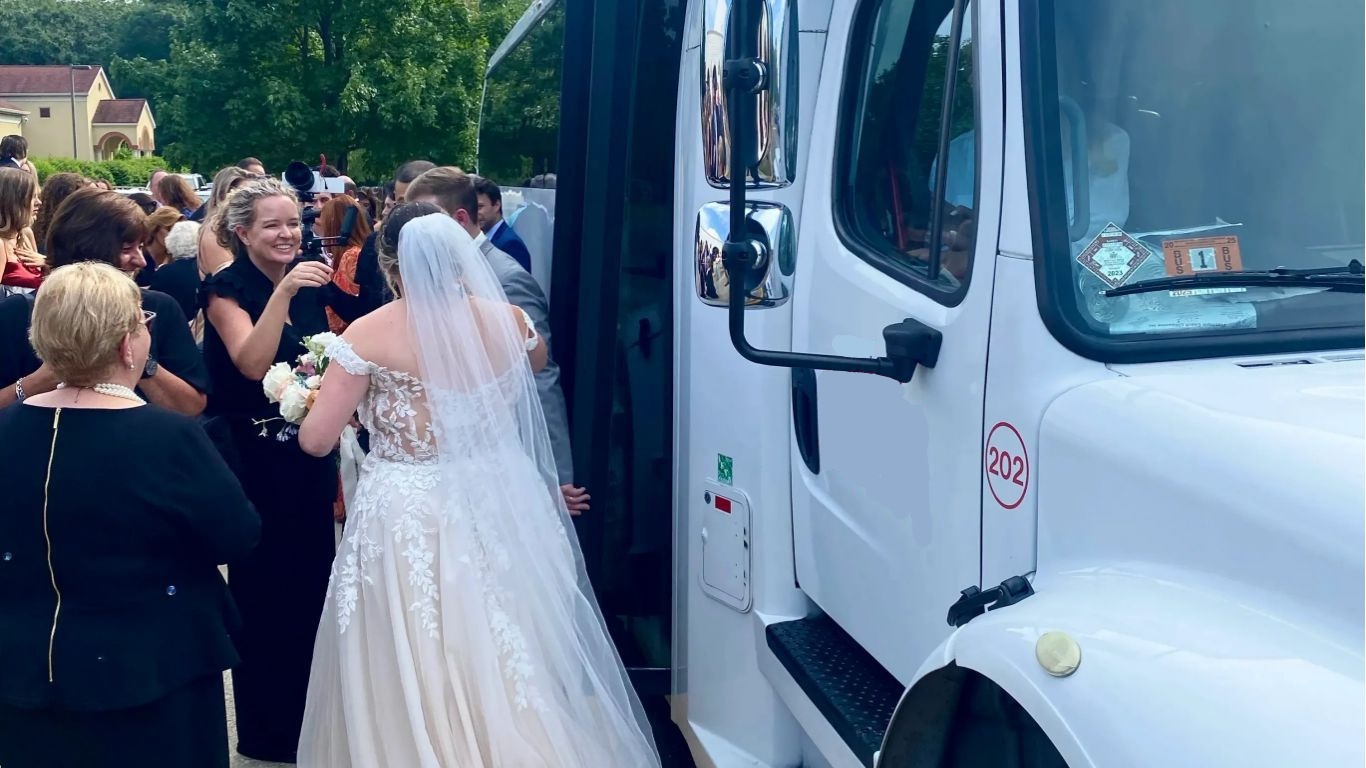 A bride in a white wedding gown prepares to board a white shuttle bus while guests gather nearby on a sunny day.
