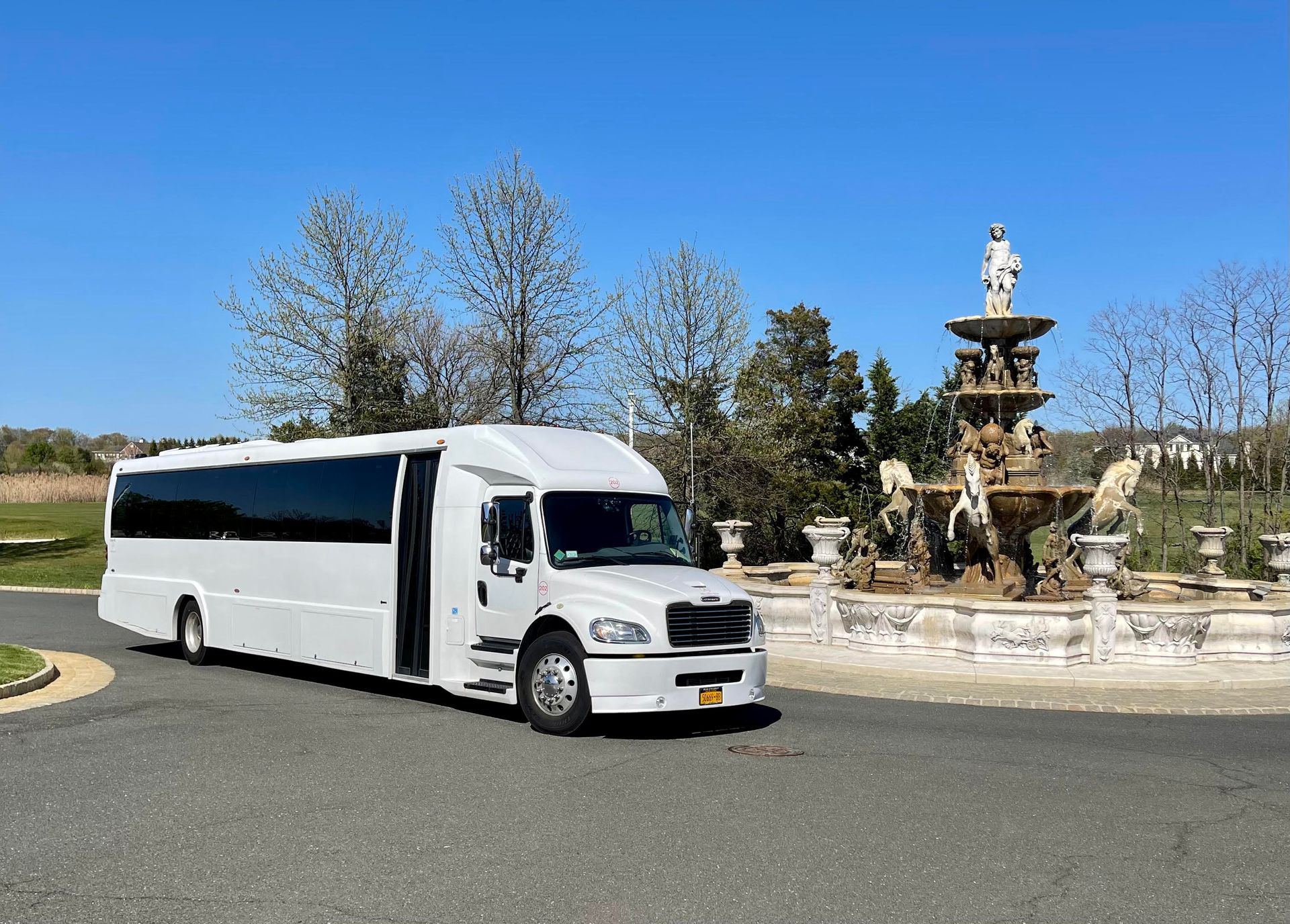 A white shuttle bus parked on a paved driveway in front of a decorative outdoor stone fountain under a clear blue sky.