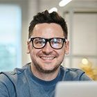 A person with dark hair and glasses smiles while sitting in a brightly lit, blurred office setting.