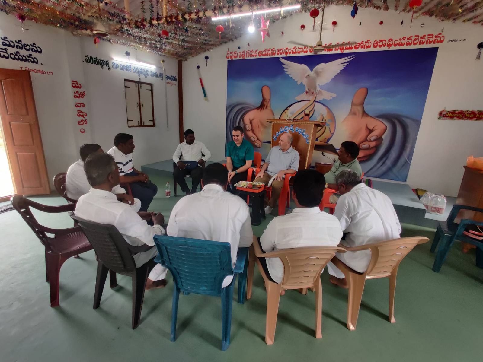A group of men are sitting in a circle in front of a painting of jesus giving a thumbs up