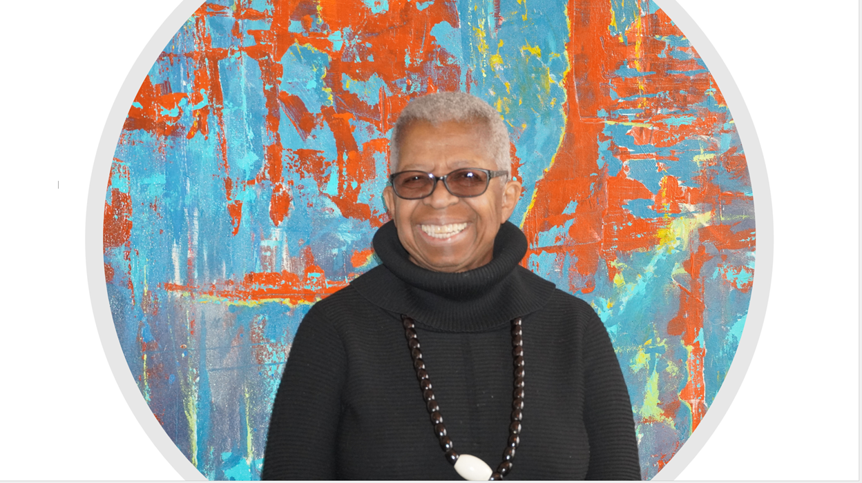 A woman wearing glasses and a black turtleneck is smiling in front of a colorful painting.