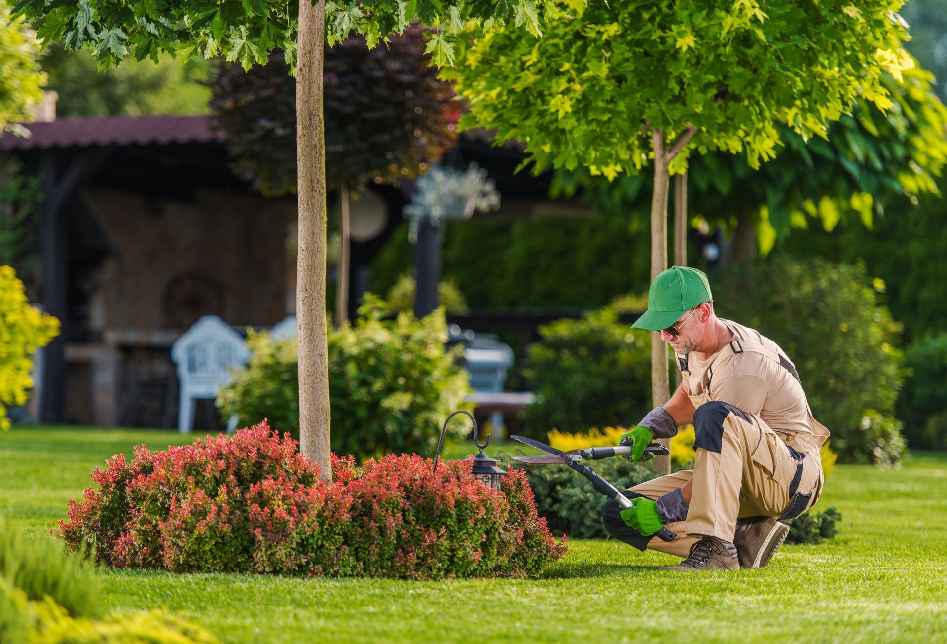 Gardener trimming bushes in a lush, green yard with a gazebo in the background.