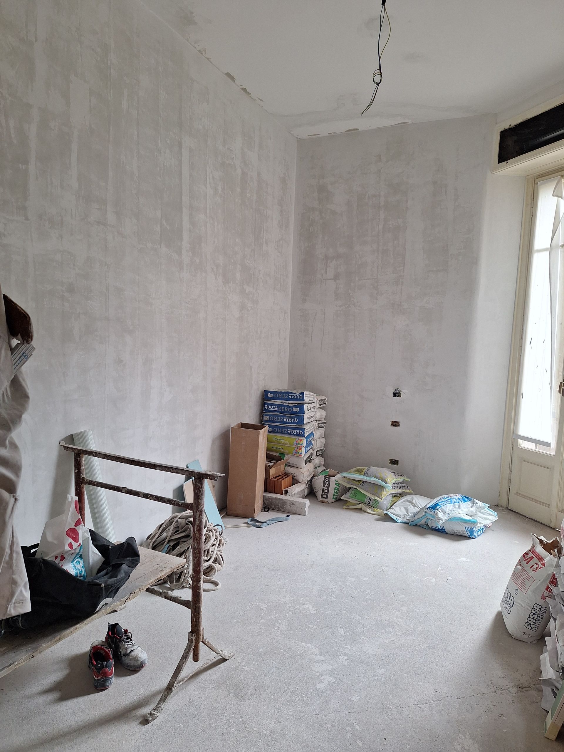 Stanza di ristrutturazione: pareti con intonaco strutturato, materiali da costruzione sul pavimento, un appendiabiti e una porta.