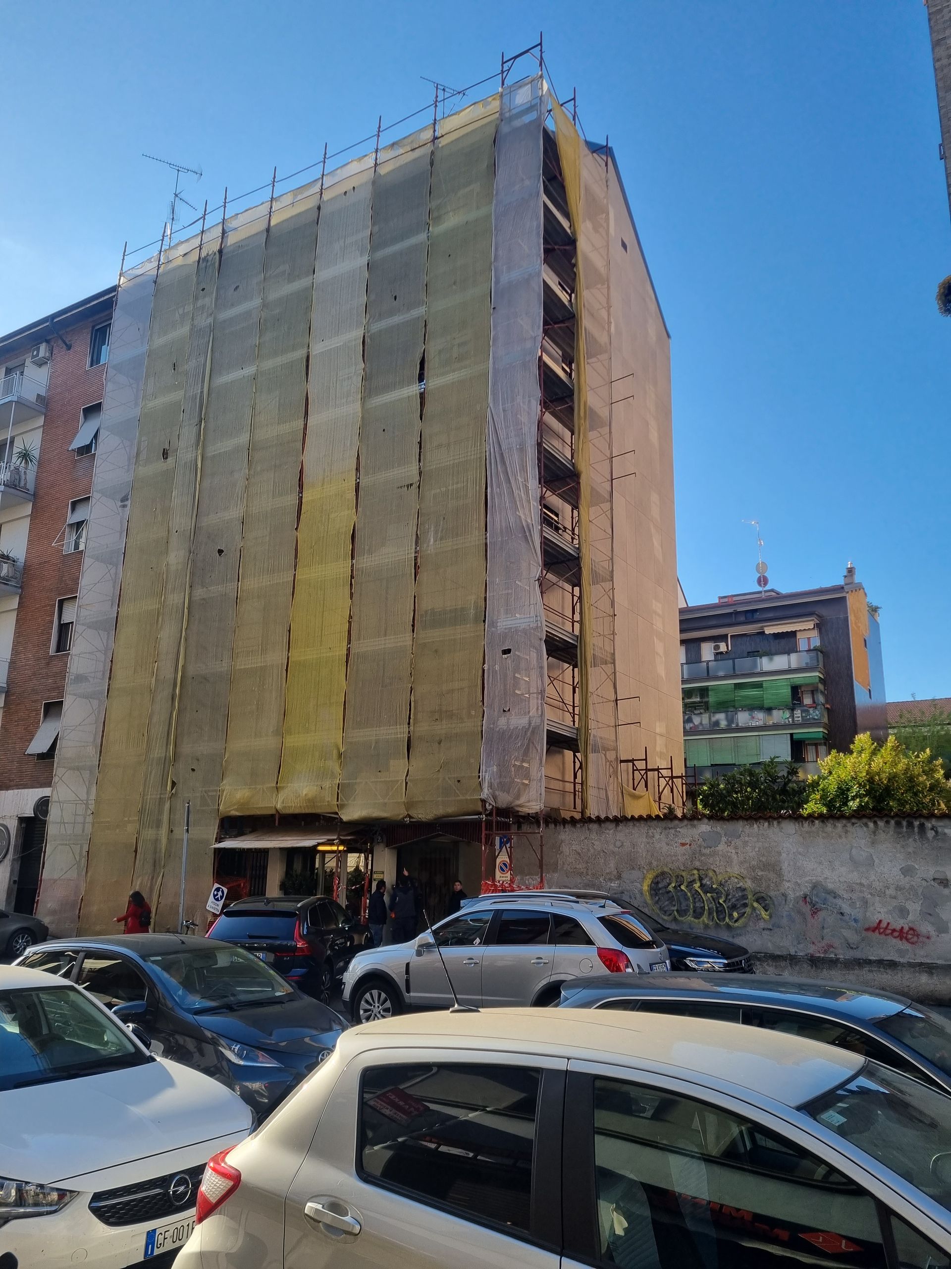 Cantiere con recinzione di sicurezza arancione, un escavatore e un operaio. Edifici sullo sfondo.