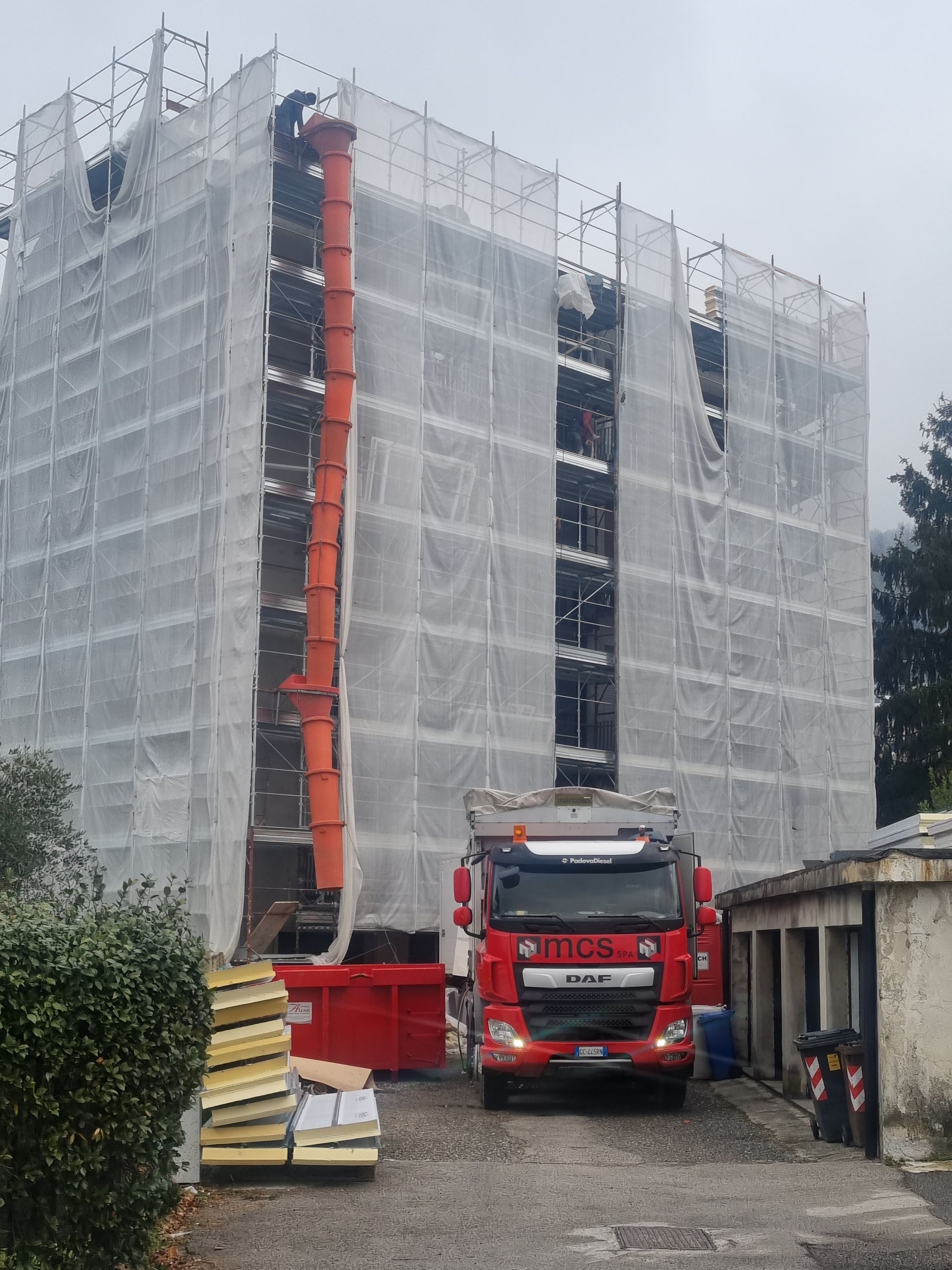 Camion rosso e cassone per rifiuti vicino a un edificio in costruzione, con uno scivolo per detriti in funzione.