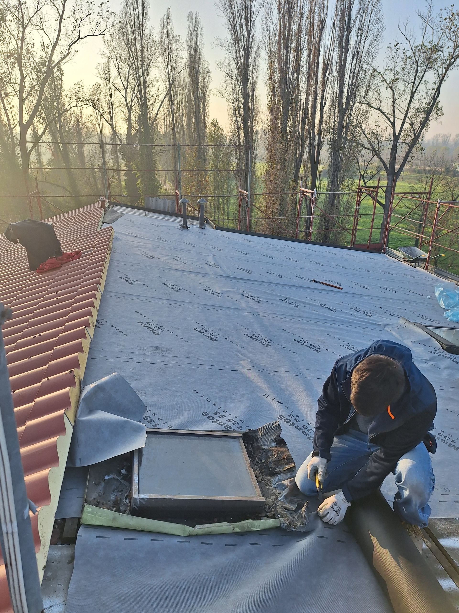 Operaio edile su un tetto, impegnato nell'installazione di materiali di copertura. Alberi e cielo sullo sfondo.