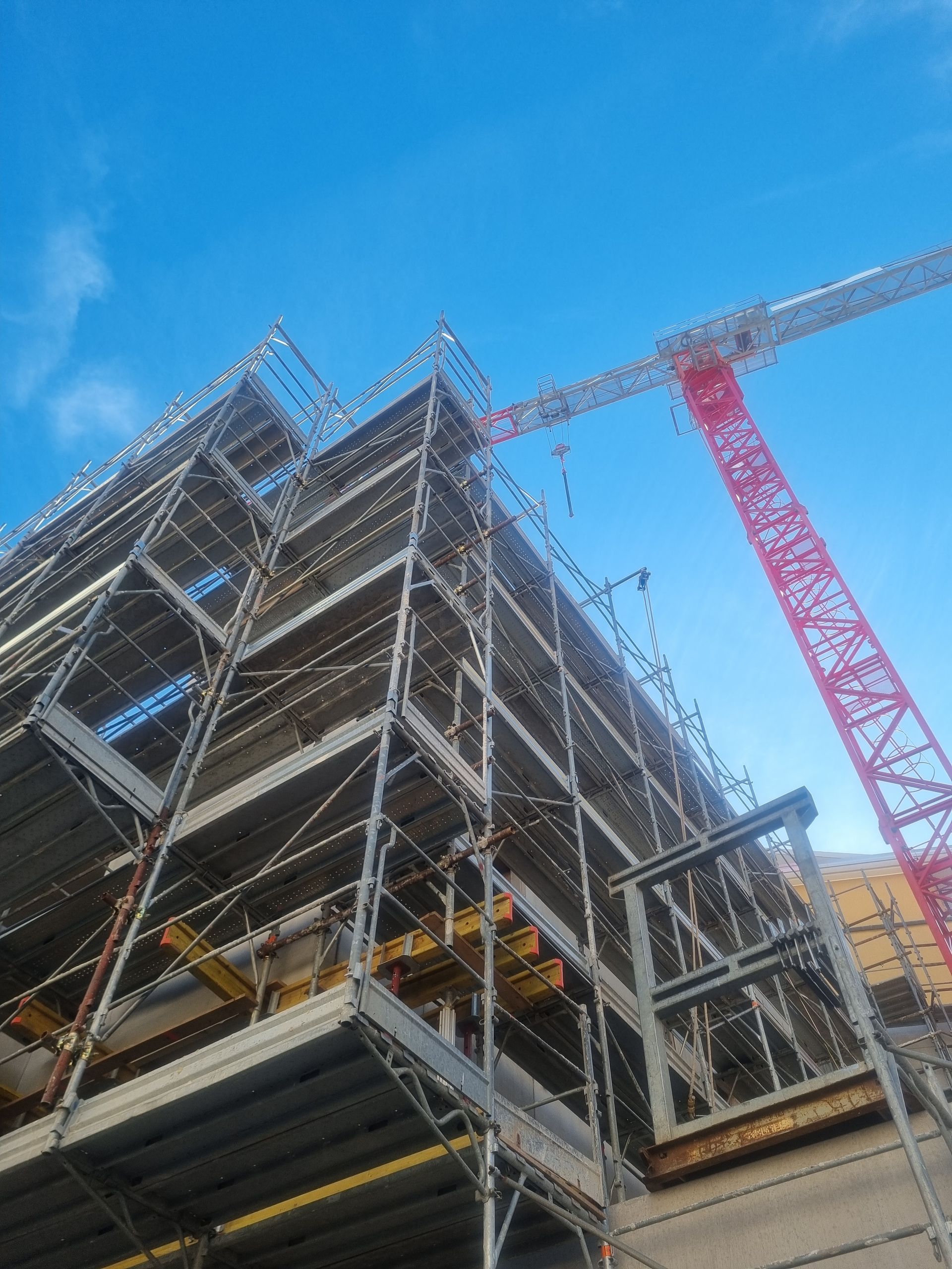 Edificio con struttura in acciaio in costruzione con una gru rossa sullo sfondo di un cielo azzurro brillante.