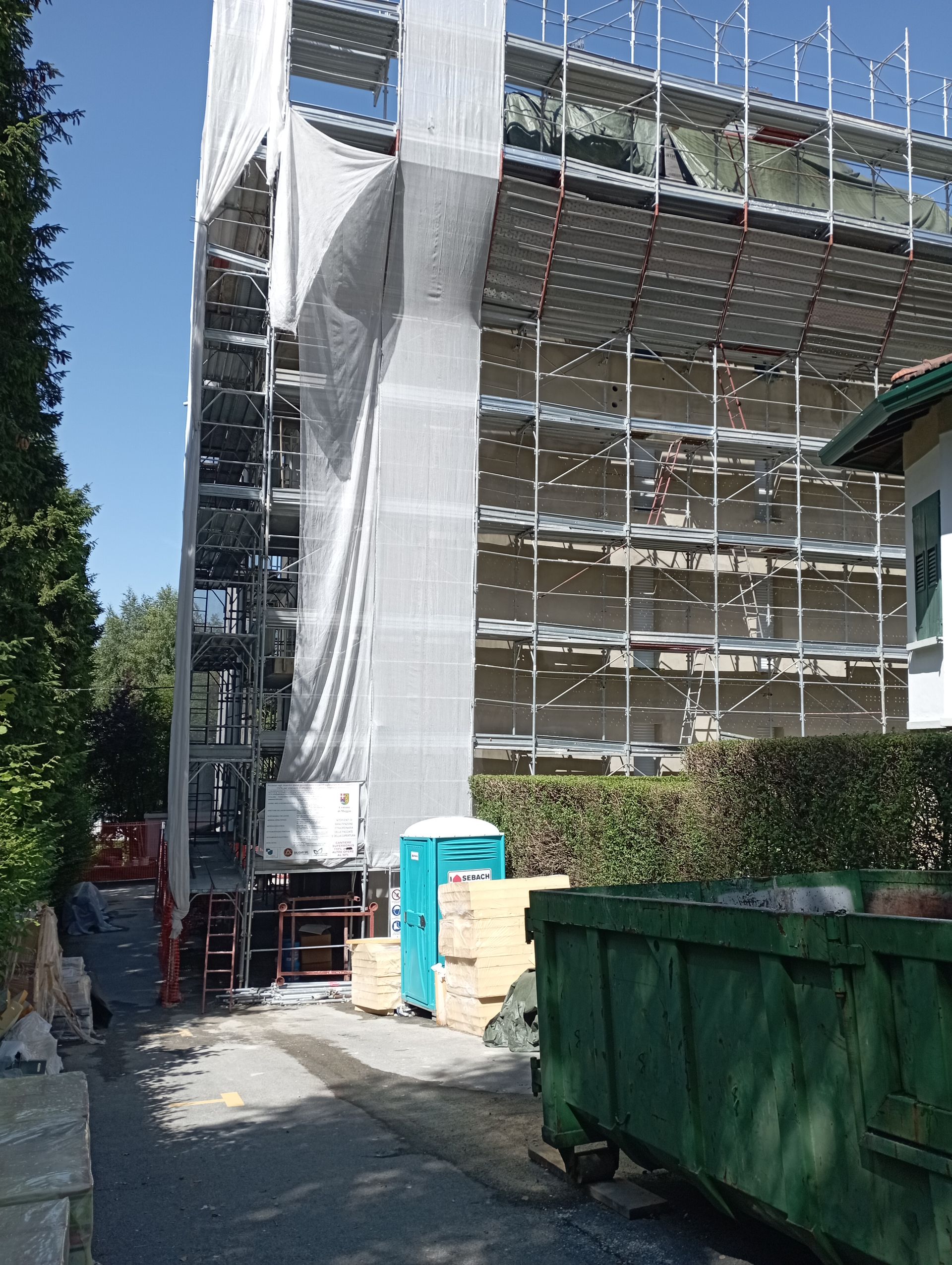 Edificio in costruzione, impalcature, reti, bagni chimici, cassonetti e alberi alla luce del giorno.