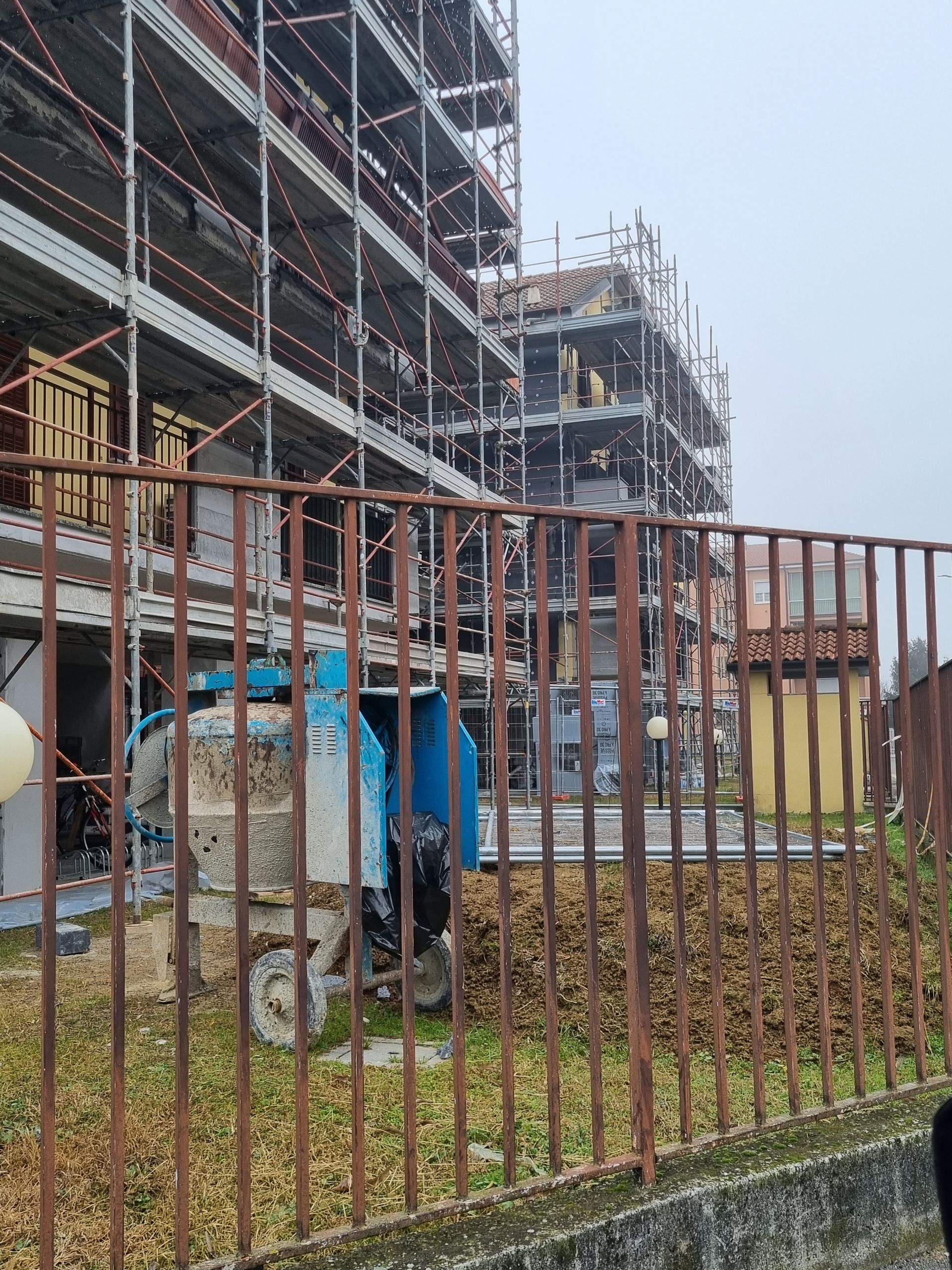 Edificio in costruzione con impalcature, una betoniera blu davanti a una recinzione marrone.