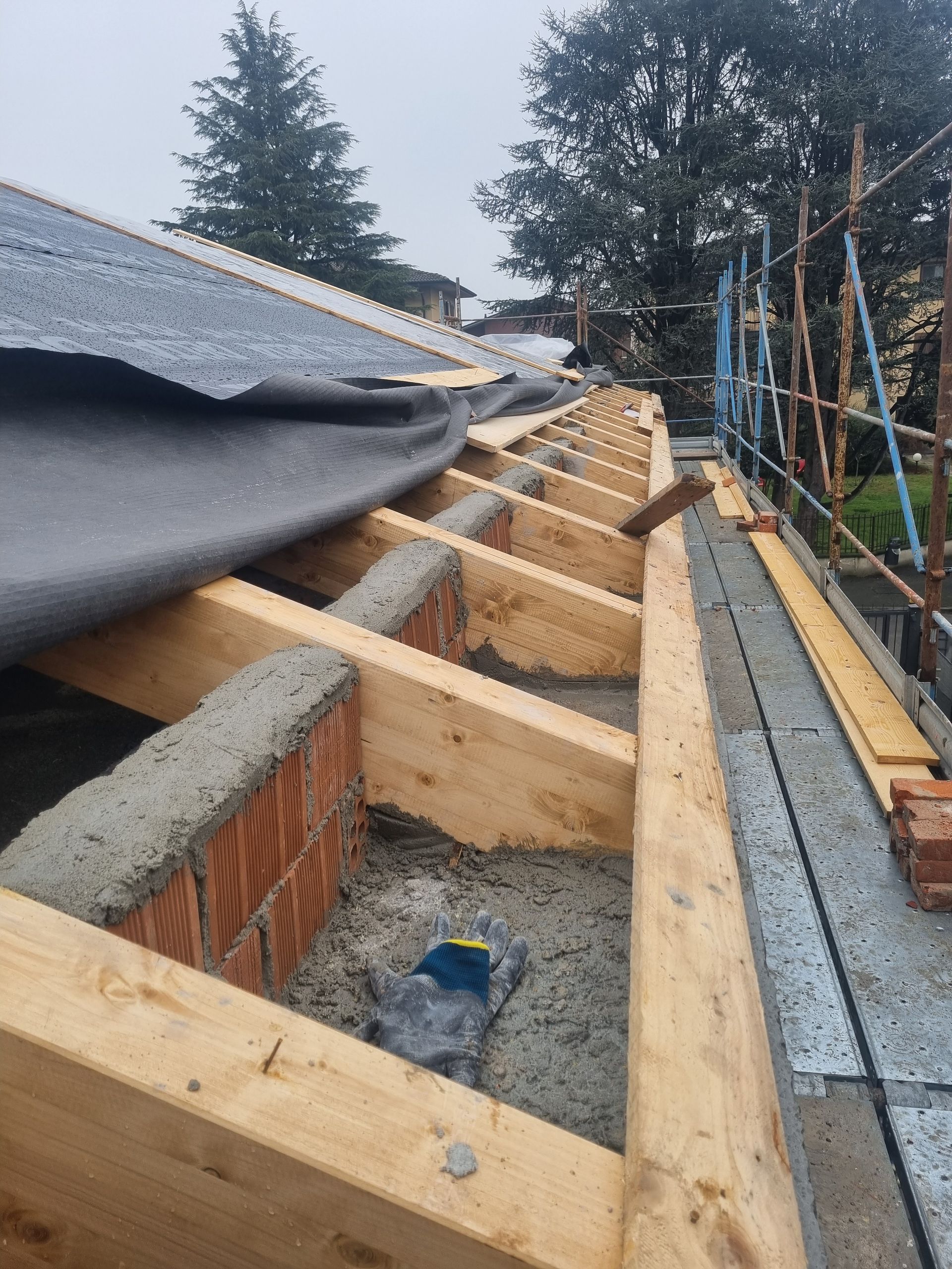Costruzione del tetto in corso: travi in ​​legno, mattoni e cemento grigio, con materiale di copertura nero parzialmente installato.
