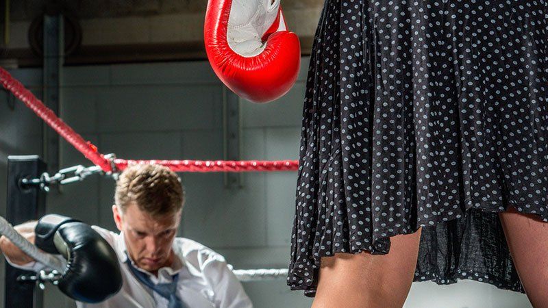 Person in dress with boxing gloves standing over a man sitting down in the corner