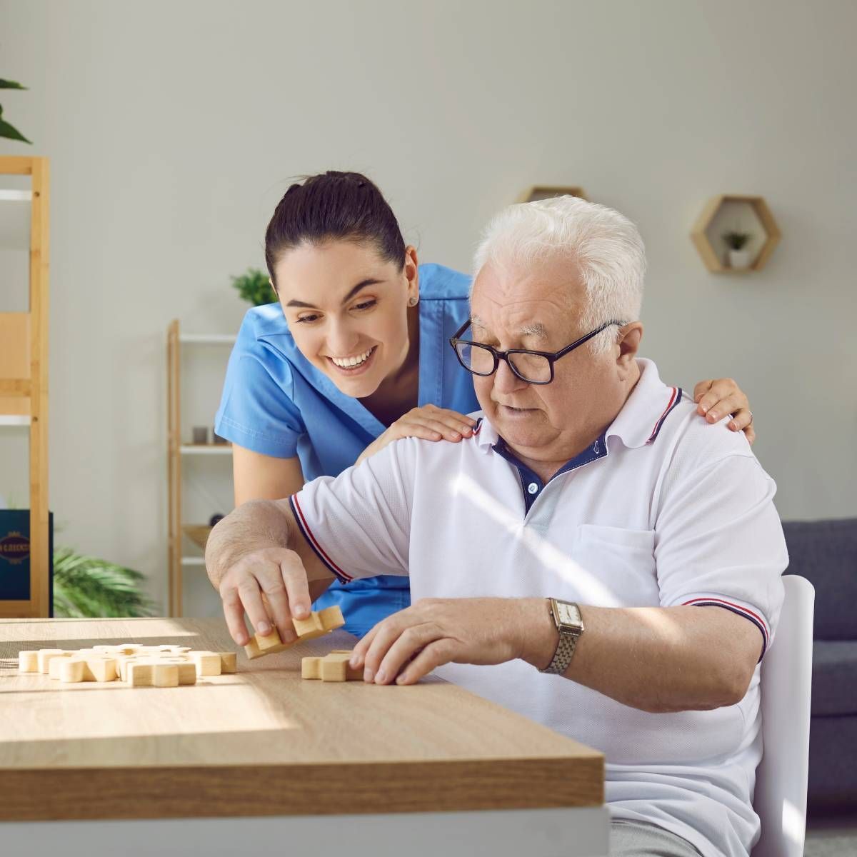 Woman in blue scrubs assists older man playing a game at a table.