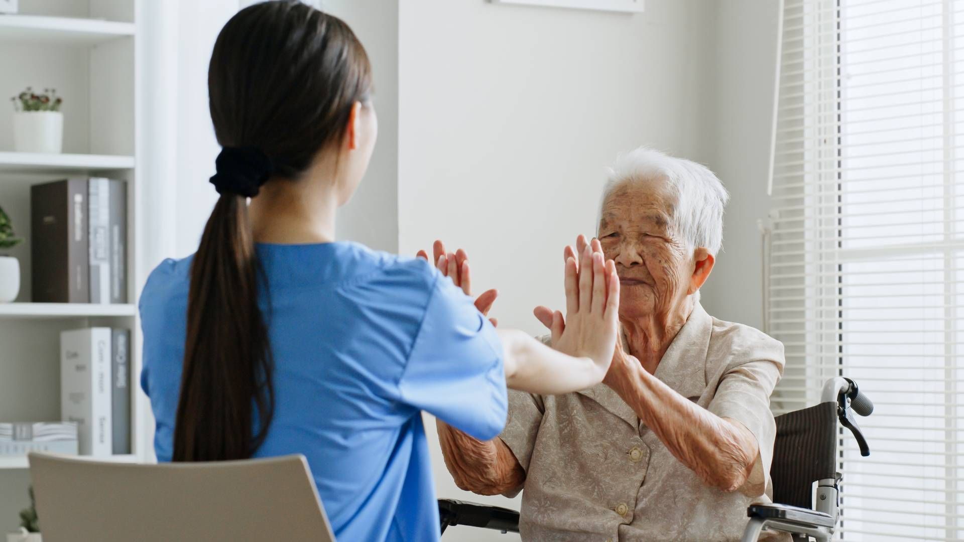 Caregiver assisting an elderly person with arm exercises in a bright room.