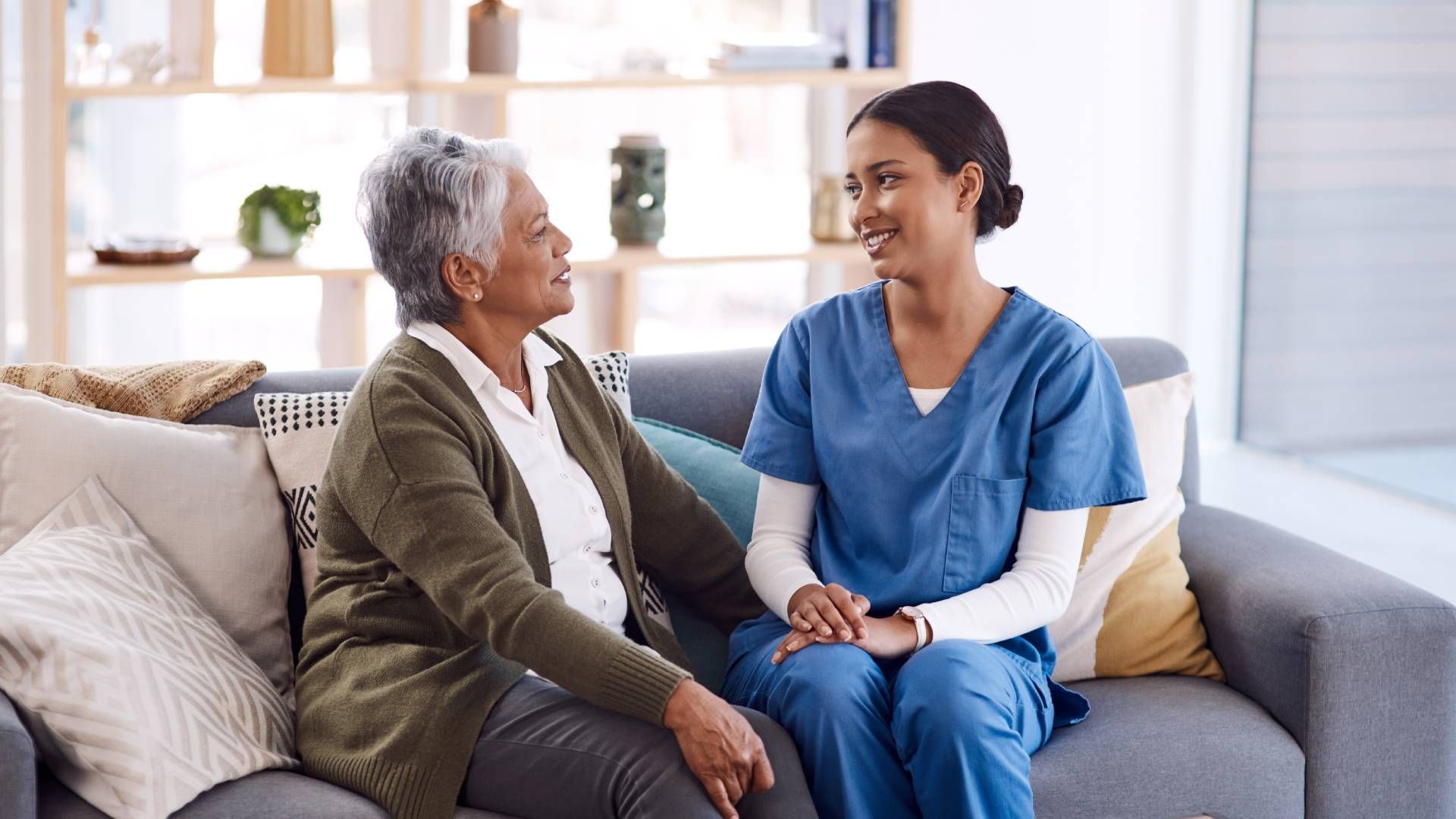 Woman in scrubs converses with an older person on a couch in a living room.