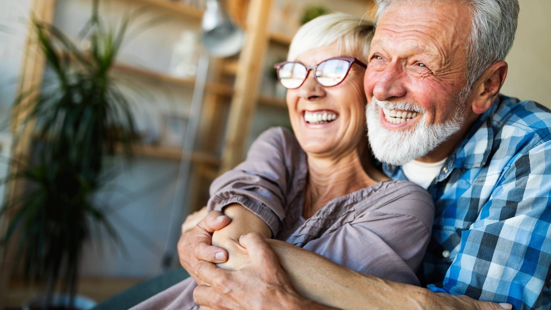 Smiling older couple embracing indoors. Man with beard, woman with glasses.