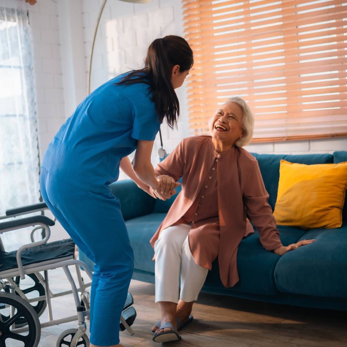 Caregiver assisting a person sitting on a sofa. The person smiles as they are helped to stand. Wheelchair nearby.