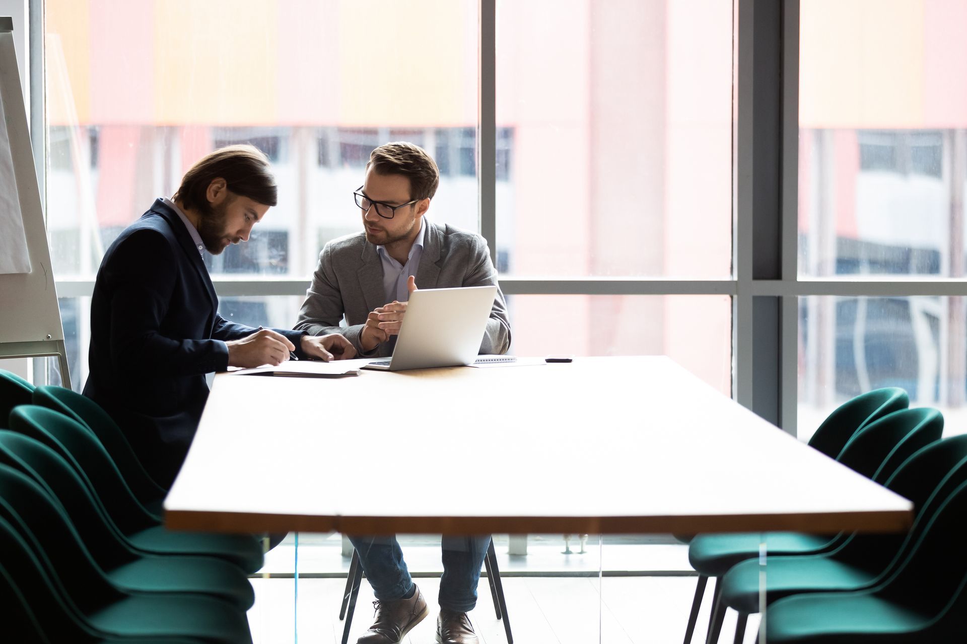 Two men are sitting at a conference table looking at a laptop.