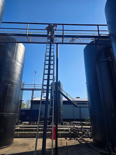A man is standing on a ladder over a bridge between two large black tanks.