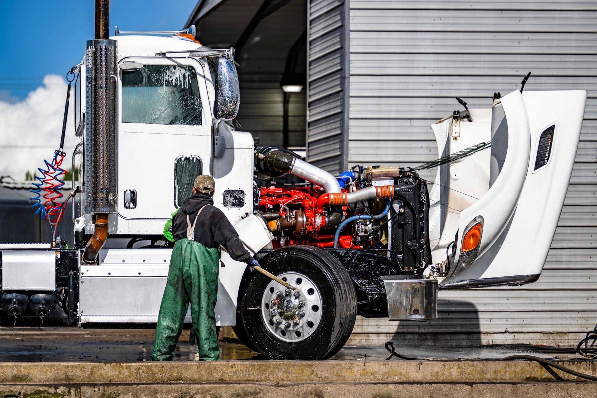 A person washes a white semi-truck with the hood open, revealing a red engine, in a garage.