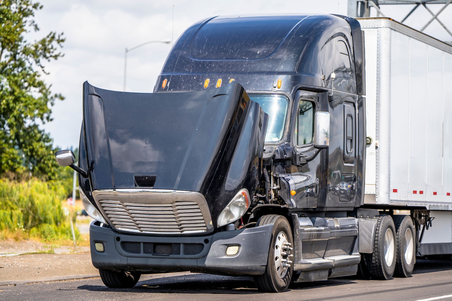 Damaged black semi-truck with open hood on the side of a road.