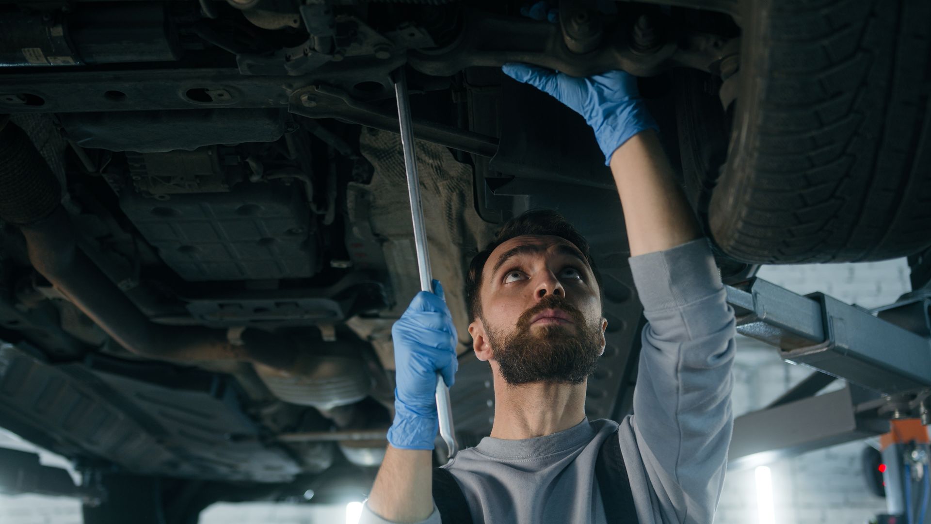 Mechanic wearing blue gloves working under a car, holding a part, in a garage.
