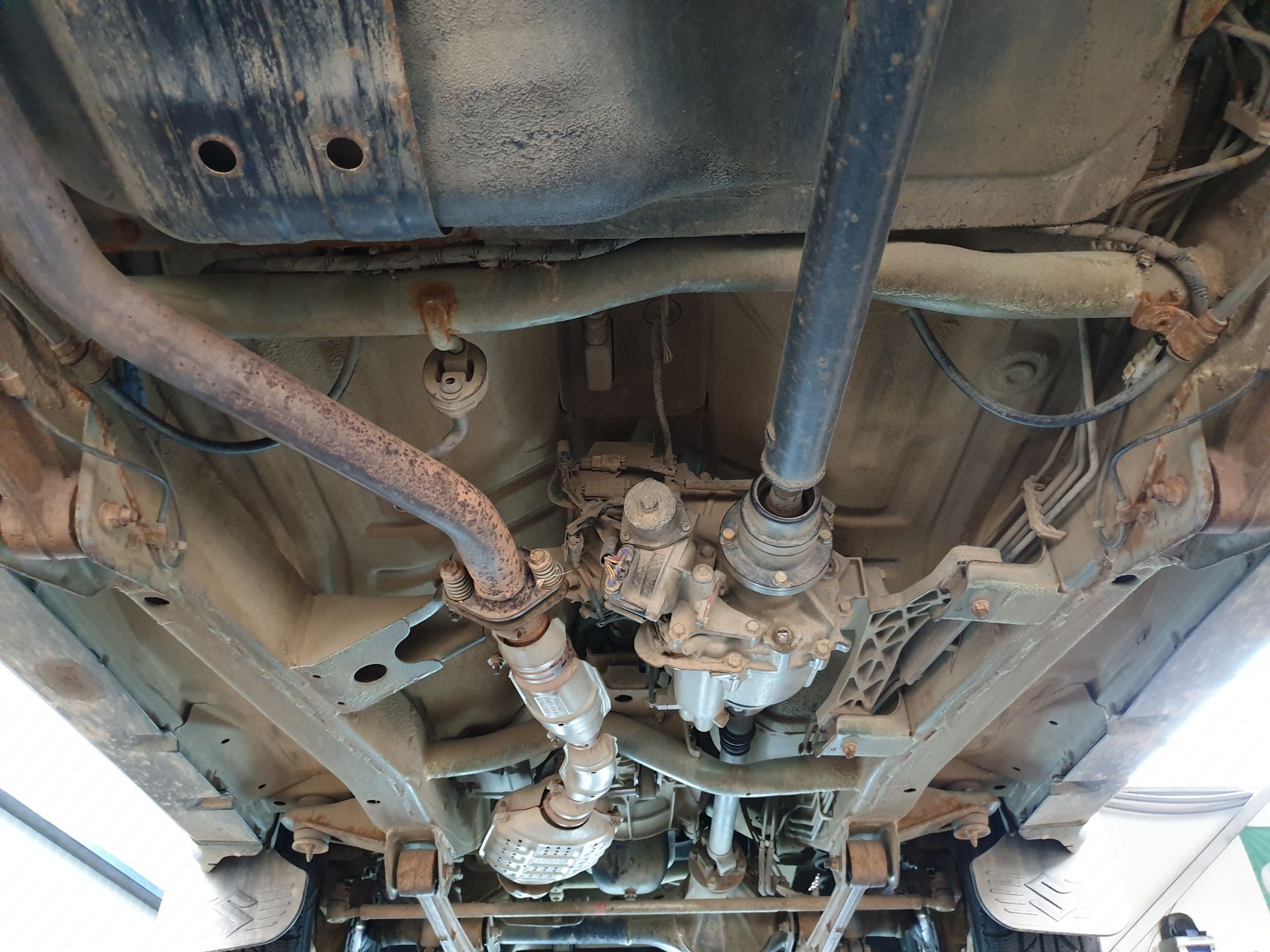 Undercarriage of a vehicle with exhaust pipe, drive shaft, transfer case, and suspension components, viewed from below.