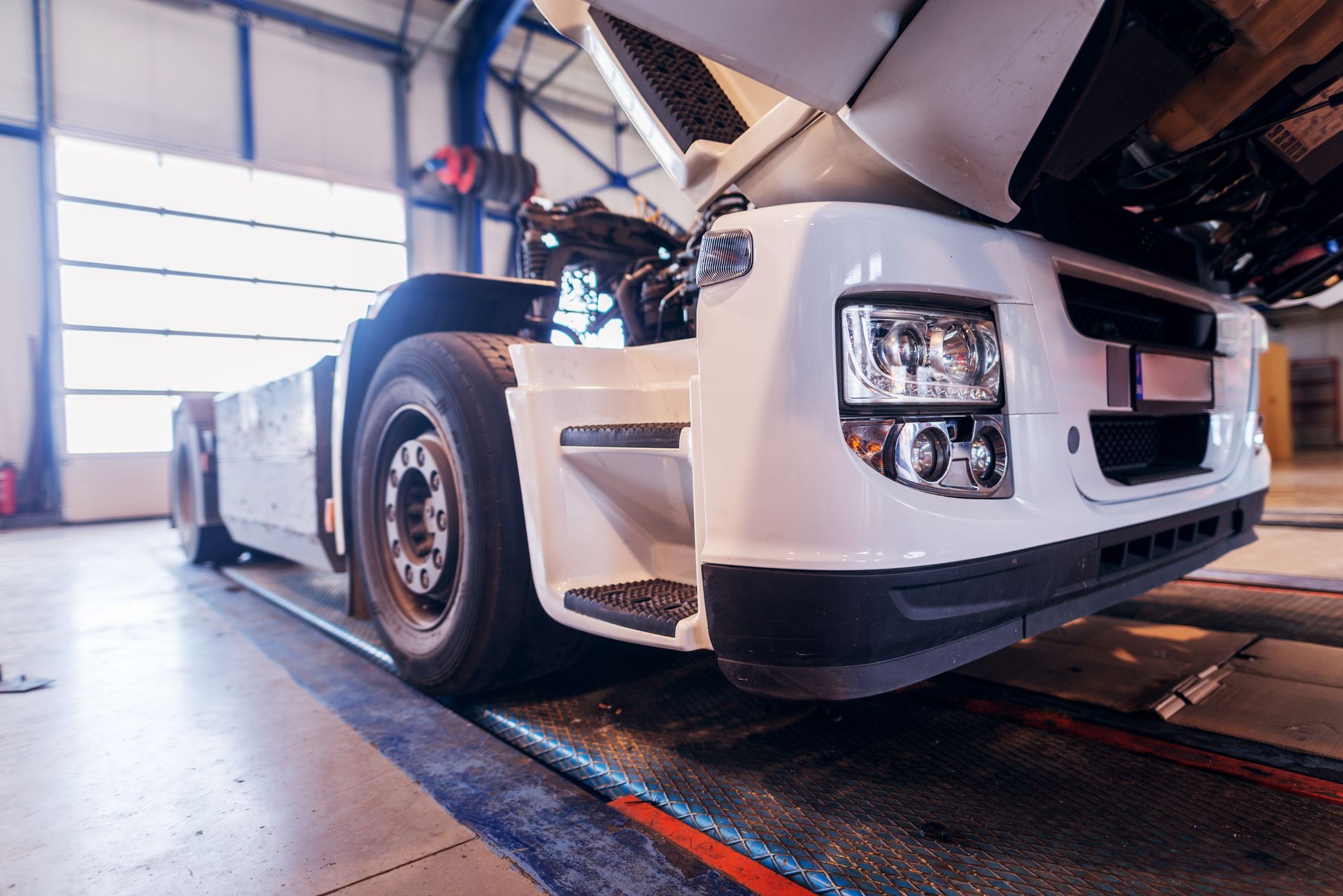 White semi-truck in a repair shop with hood open, near a window, ready for maintenance.